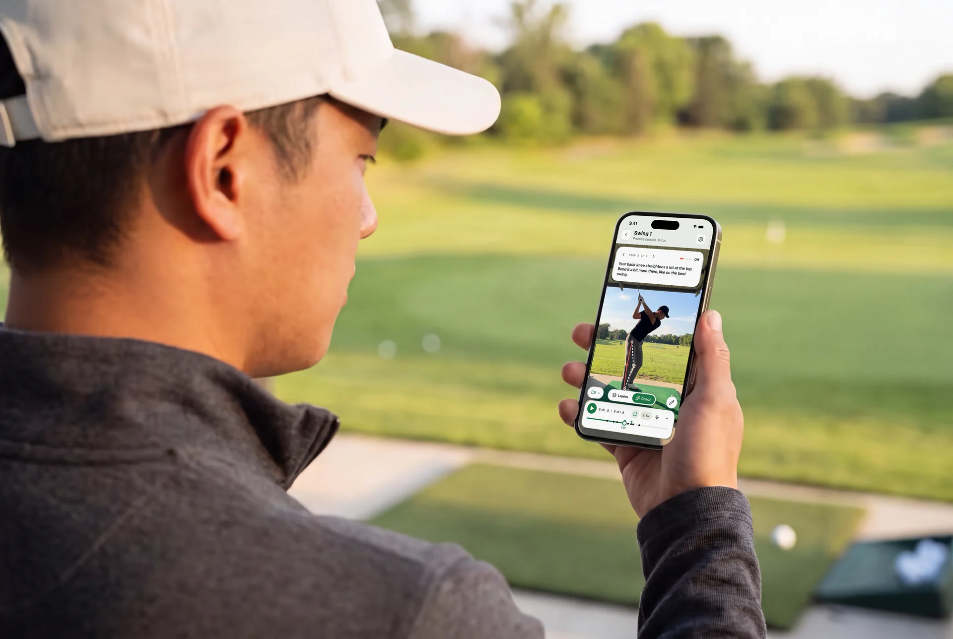 A golfer at the driving range looks at an iPhone. The screen shows coach step 2: bend the back knee a bit more at the top.